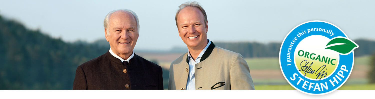 Two men stand outdoors with a circular logo featuring the word “Organic” and the name “Stefan Hipp” in the foreground.