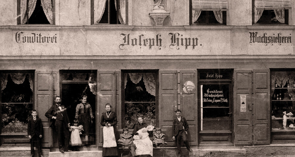 A group of seven people, dressed in early 20th-century attire, stand and sit outside a two-story building with signage in a European language.