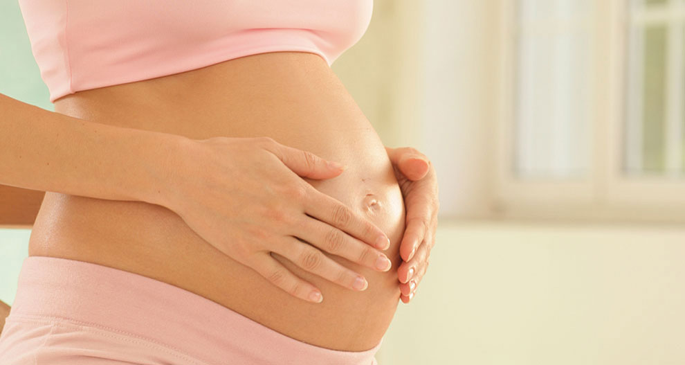 A pregnant woman in pink clothing, with her hands gently placed on her belly, is seated indoors.