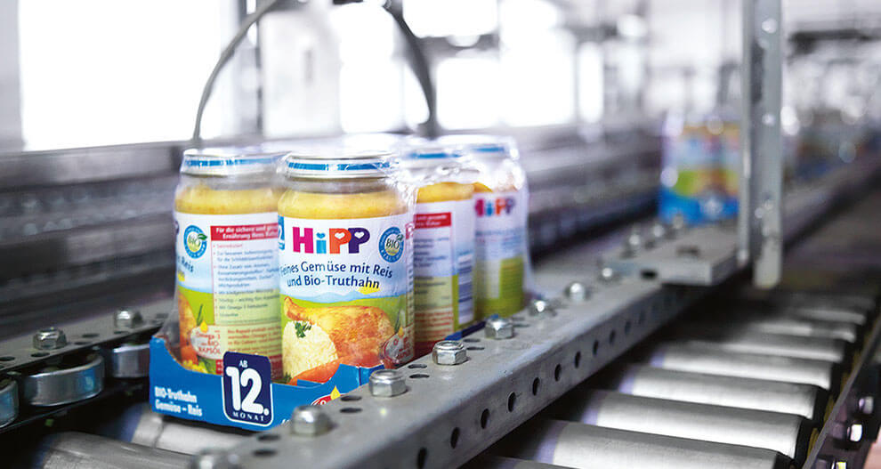 A conveyor belt in a factory setting transports jars of baby food with a label that includes the word "HiPP" and a picture of a food item.