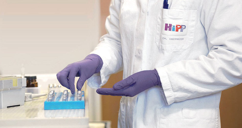 A person in a white lab coat with the logo "HiPP" and the name "CH. OBERMAIER" is wearing purple gloves and handling a blue rack with small containers.