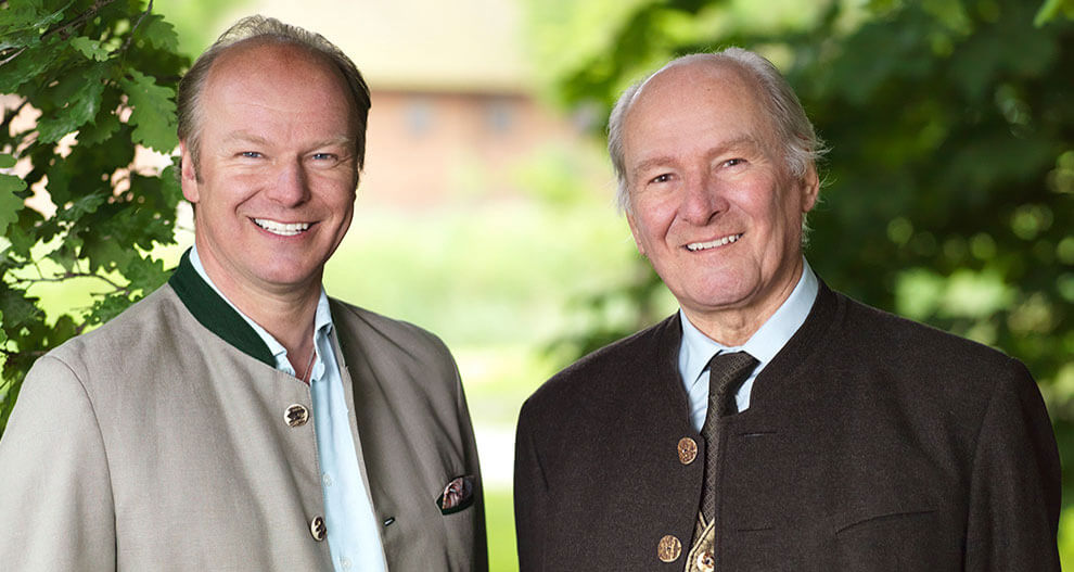 Two men, one in a light-colored jacket and the other in a dark-colored jacket, stand side by side outdoors.