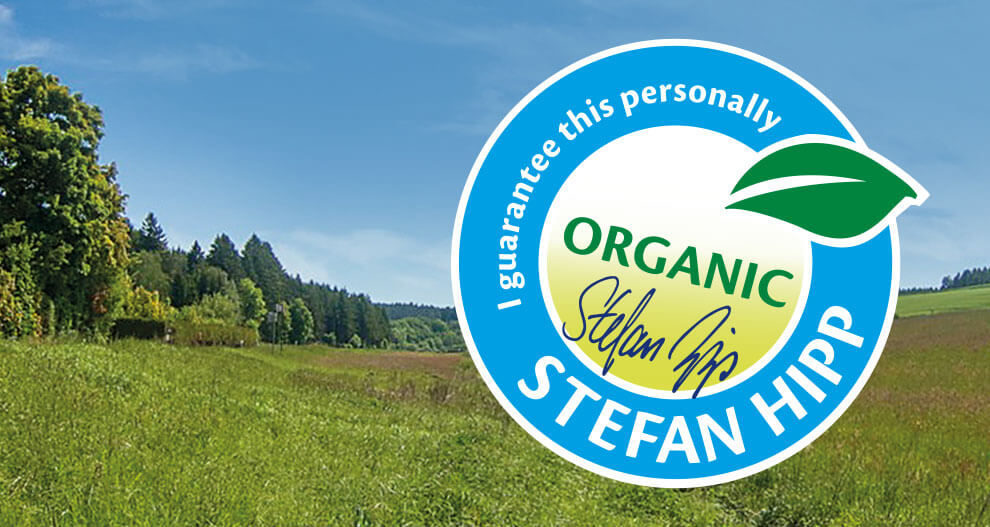 A landscape with green grass and trees is overlaid with a blue circular graphic containing the word “ORGANIC” and a signature.