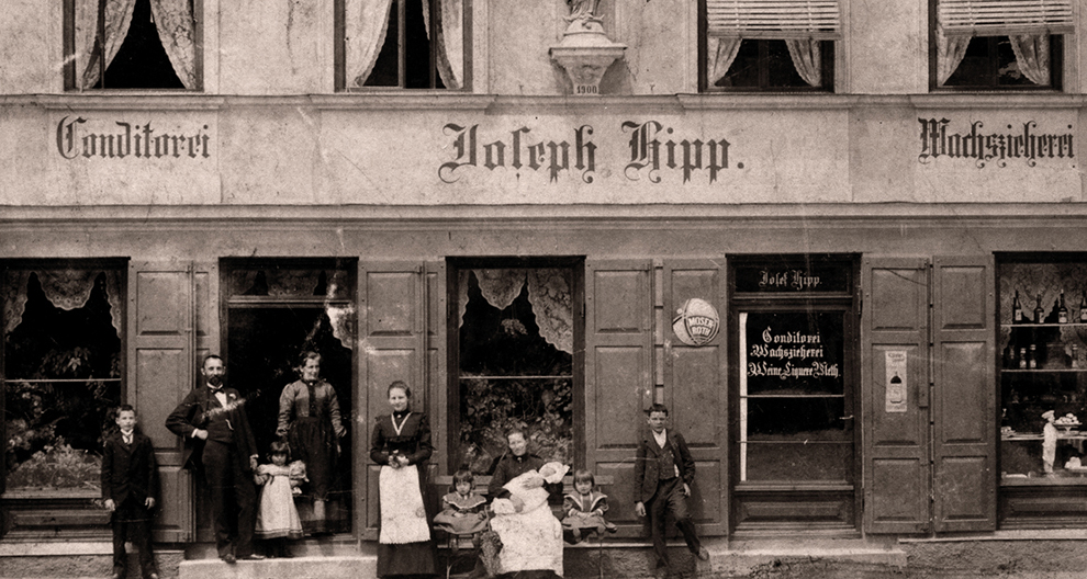 A group of people, including adults and children, stand and sit outside a two-story building with signage in a European setting.