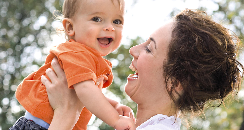 A woman in a white shirt holds a baby in an orange shirt, both looking at each other with a joyful expression.