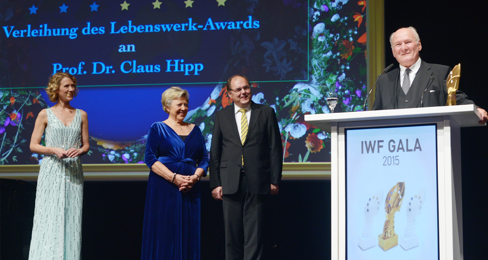 Four individuals stand on a stage with a backdrop displaying German text and a floral design, and a podium to the right displays the words "IWF GALA 2015" and an image of a trophy.