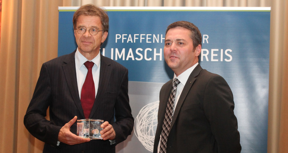 Two men in suits stand in front of a blue banner with white text, one holding a transparent award.