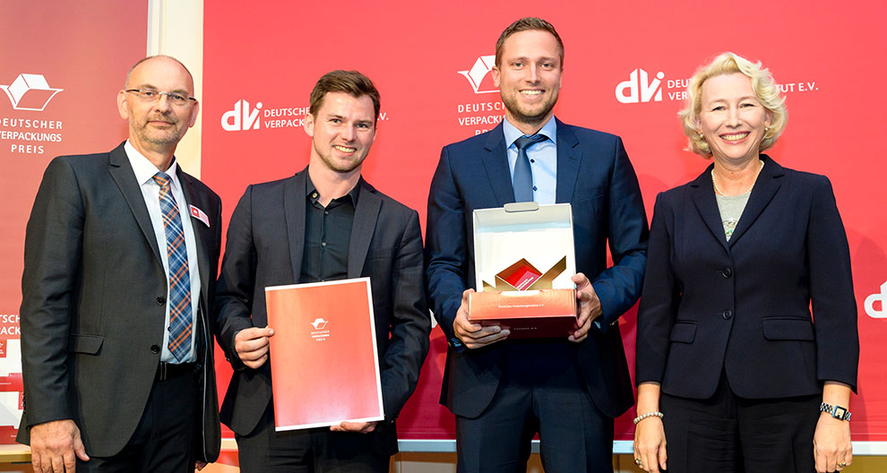 Four individuals stand in front of a red backdrop with logos and text, two of whom hold awards.