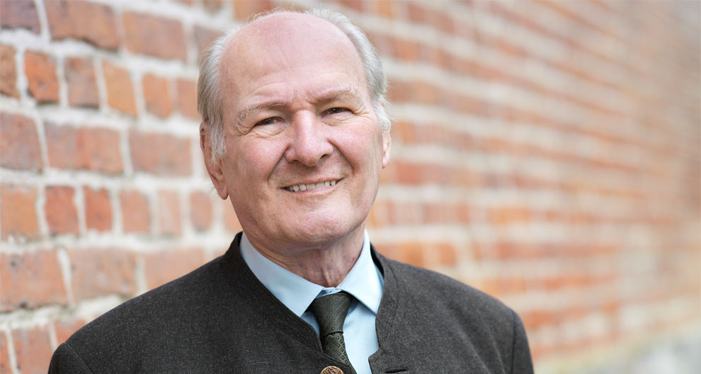 An elderly man with a bald head and a gentle smile stands in front of a brick wall, wearing a dark suit with a light blue shirt and a green tie.