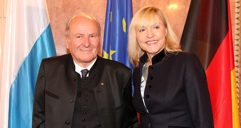 Two individuals stand side by side in front of a backdrop of flags.
