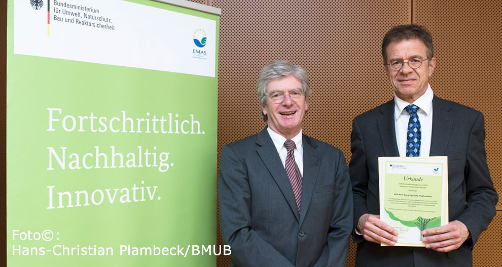 Two men in suits stand next to a green banner with German text and logos, one of whom holds a certificate.